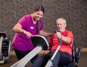 Senior male working out on a recumbent bike while an attentive staff member gives guidance