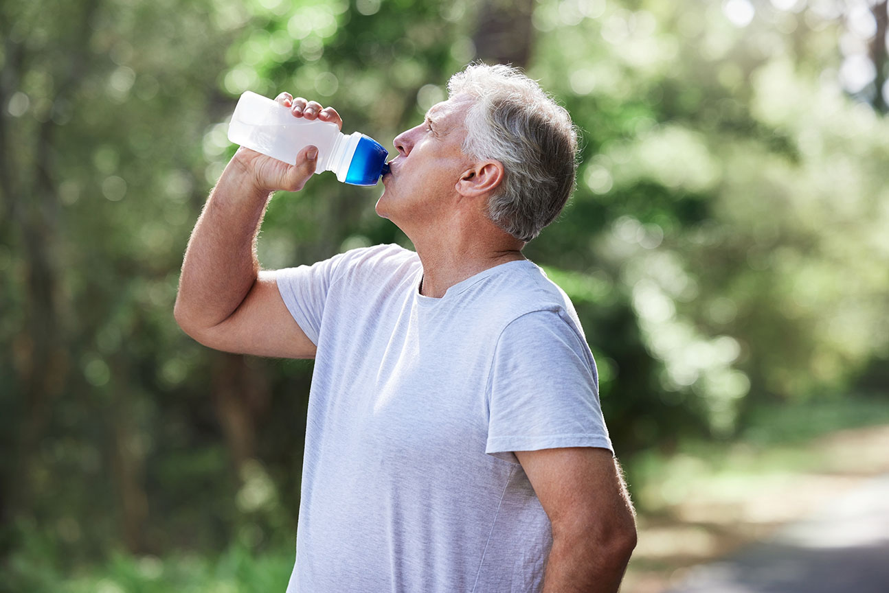 A senior man is exercising outdoors and drinking water after a run while also wearing a light-colored t-shirt to not overheat from the sun.